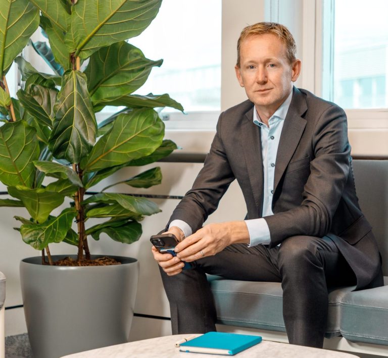 Joe O’Malley, Managing Partner, seated in a modern office space beside a large potted plant, holding a remote control with a notebook on the table in front of him.