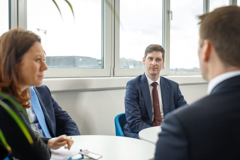 Four Hayes Solicitors LLP professionals seated around a table in a bright meeting room, engaged in conversation with large windows in the background.