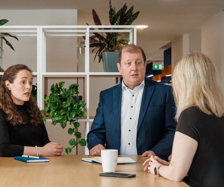 Three colleagues at Hayes Solicitors LLP engaged in a conversation around a table in a modern office space, with plants and shelving units in the background.